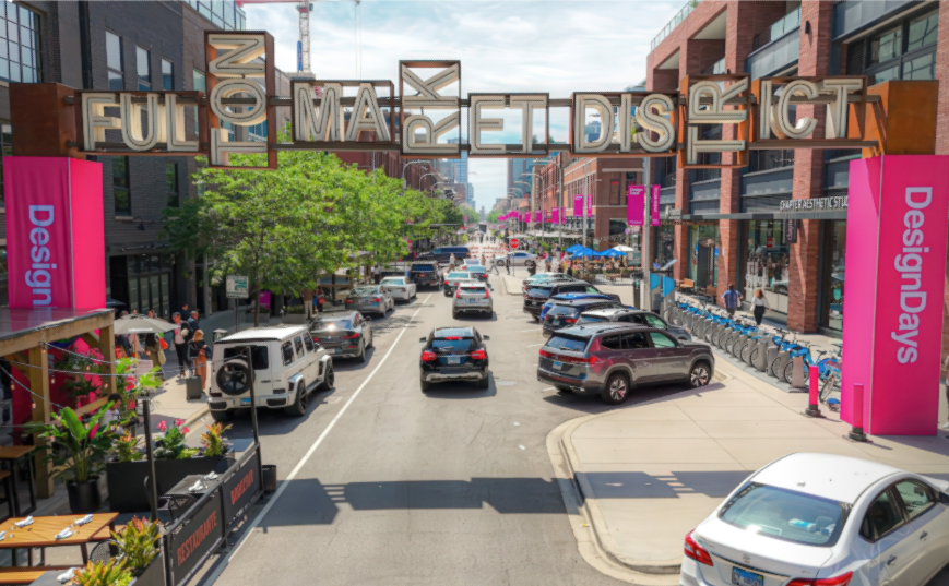 Elevated view of a lively street in Chicago’s Fulton Market District during Design Days, featuring modern buildings, outdoor dining areas, pedestrians, bicycles, and a large overhead sign reading “FULTON MARKET DISTRICT.” Bright pink banners line the street, and the scene includes cars and a white SUV, capturing the vibrant atmosphere of the event.